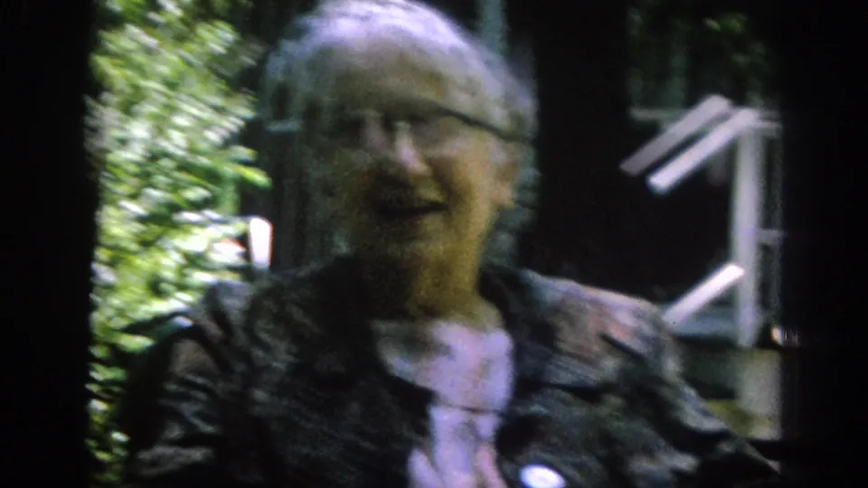 In the Blainsport home-movie scene, an older woman in glasses and patterned outerwear faces the camera near porch details and foliage.
