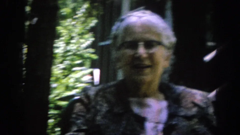 The older woman remains framed near the porch and foliage, with soft focus and dark edges from the 8mm home-movie scan.