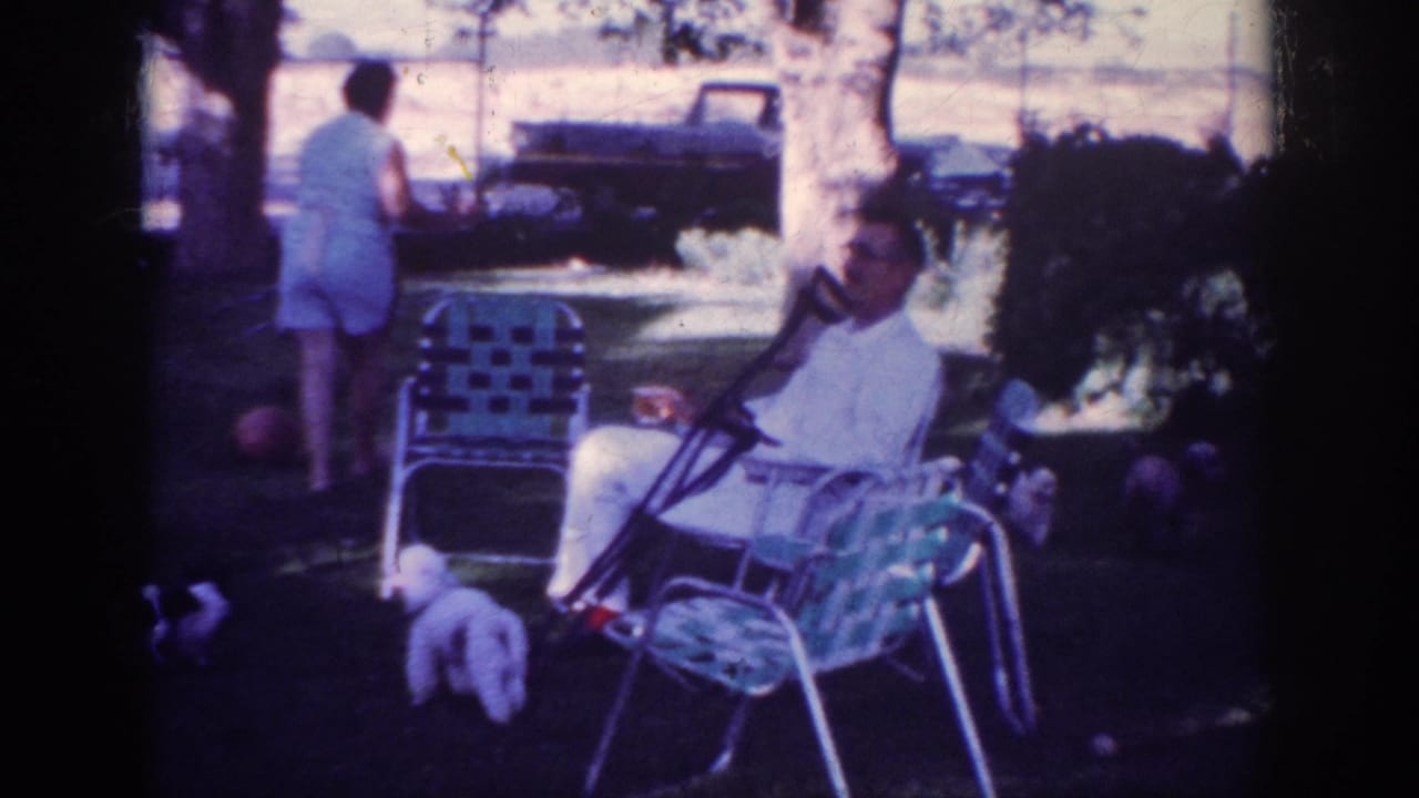 1962 Blainsport home-movie frame showing a man seated in a checkered lawn chair, a woman walking in the yard, small white dogs, trees, and a dark truck.