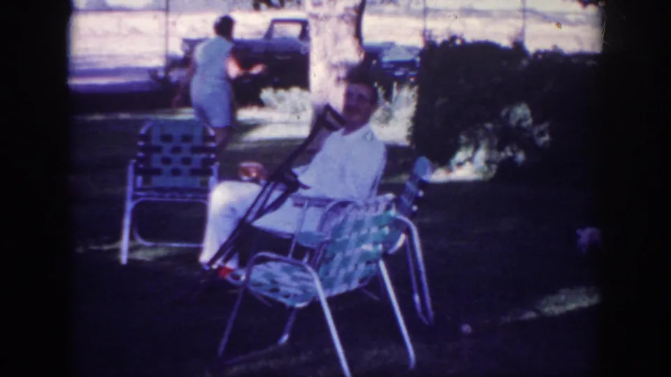1962 Blainsport yard scene with a man in a white shirt seated on a lawn chair, a woman walking near a parked truck, and small dogs nearby.