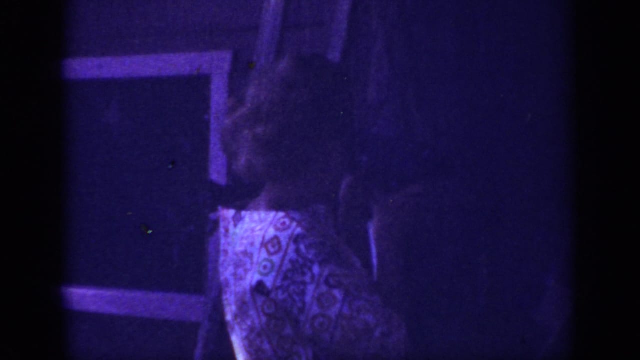 Adult woman in a patterned dress appears indoors in the 1962 Blainsport home-movie footage, partly obscured by shadow and grain.