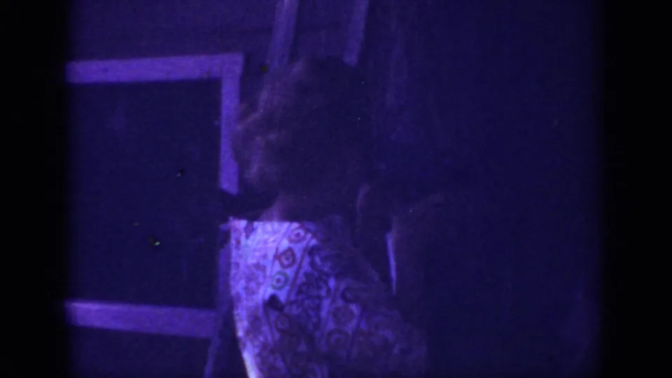Adult woman in a patterned dress appears indoors in the 1962 Blainsport home-movie footage, partly obscured by shadow and grain.