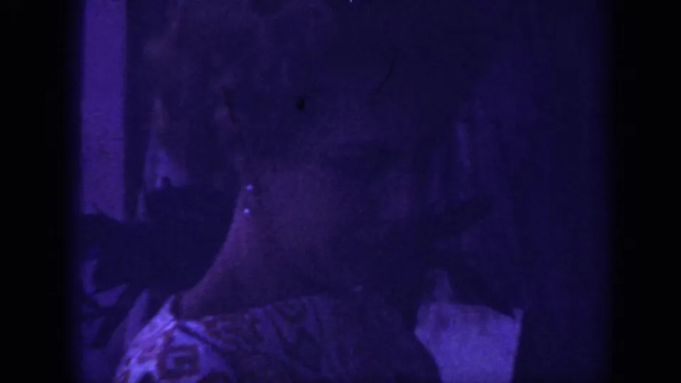 1962 Blainsport home-movie frame showing a woman close to the camera in a patterned dress, with heavy purple cast and dark gate edges.