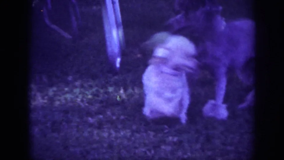 1962 Blainsport home-movie frame with a small white dog close to a bicycle wheel or metal yard object, filmed low on grass.