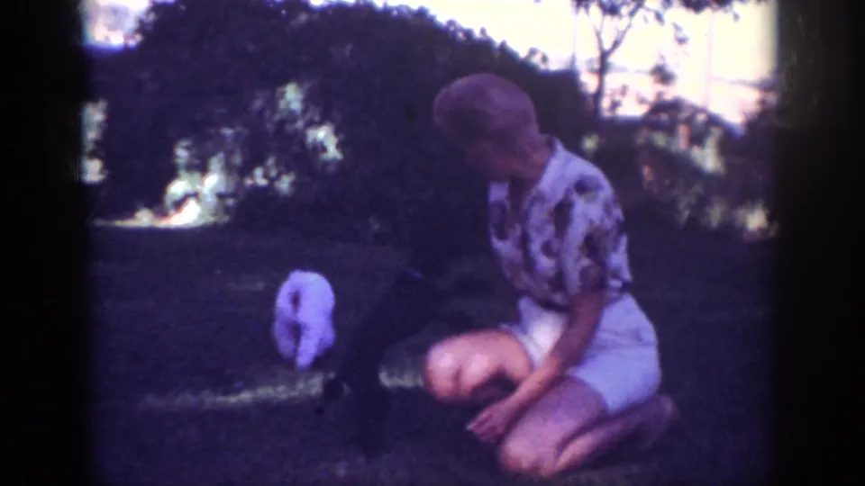 In a wide backyard view, a woman kneels beside a small white poodle on the grass, with trees and shrubs behind them.