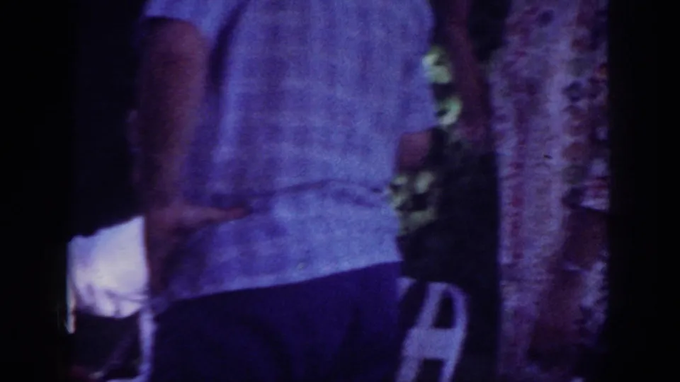 1962 Blainsport home-movie frame showing an adult in a light checked shirt near a tree at an outdoor gathering.