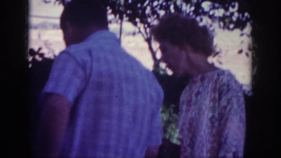 1962 Blainsport backyard frame with a man in a light shirt and a woman in a patterned dress standing under trees.