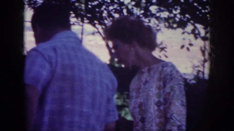In metadata-supported 1962 Blainsport, Pennsylvania footage, two adults stand near flowerbeds in a shaded backyard.