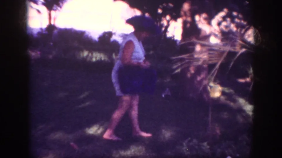 A barefoot woman in a light sleeveless dress walks through the Blainsport garden carrying a dark bag in grainy 1962 8mm footage.
