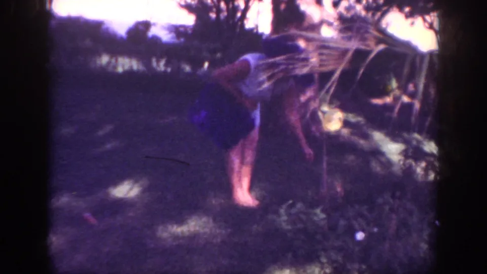 The woman bends near tall garden plants in the dim Blainsport yard, with soft focus and purple color shift from the 8mm scan.