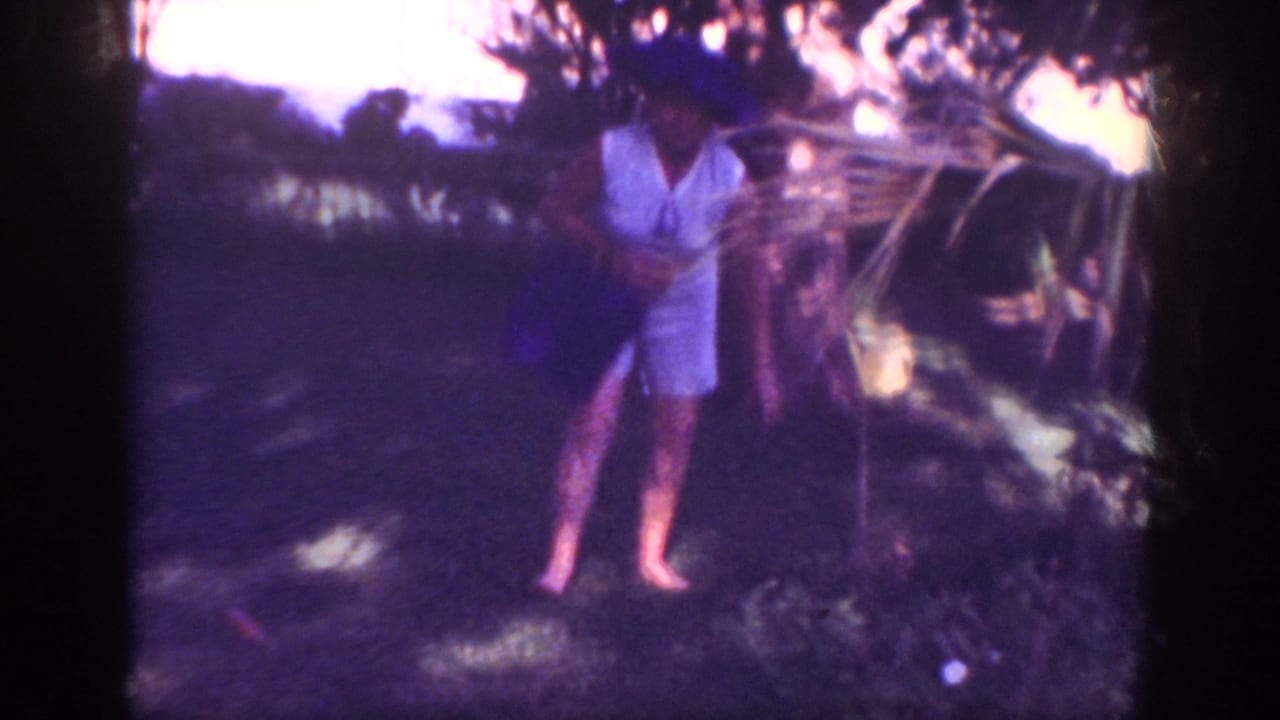 1962 Blainsport home-movie frame showing a woman in a pale sleeveless outfit and hat standing barefoot in a shaded garden.