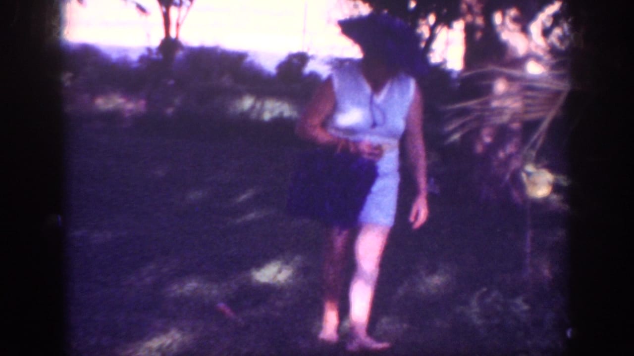 Woman walking barefoot across shaded grass while carrying a dark handbag.