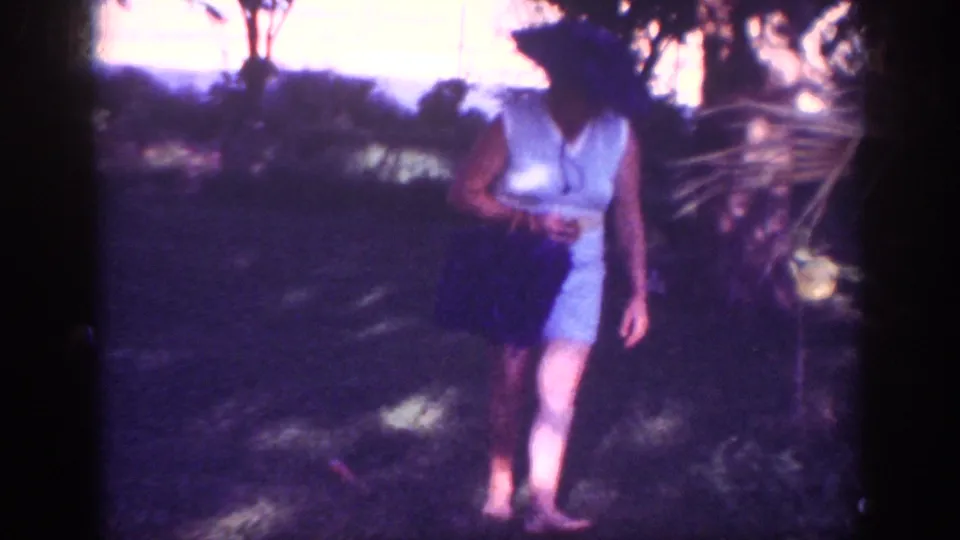 In this 1962 Blainsport garden frame, the woman walks barefoot through shade with a dark handbag beside shrubs.