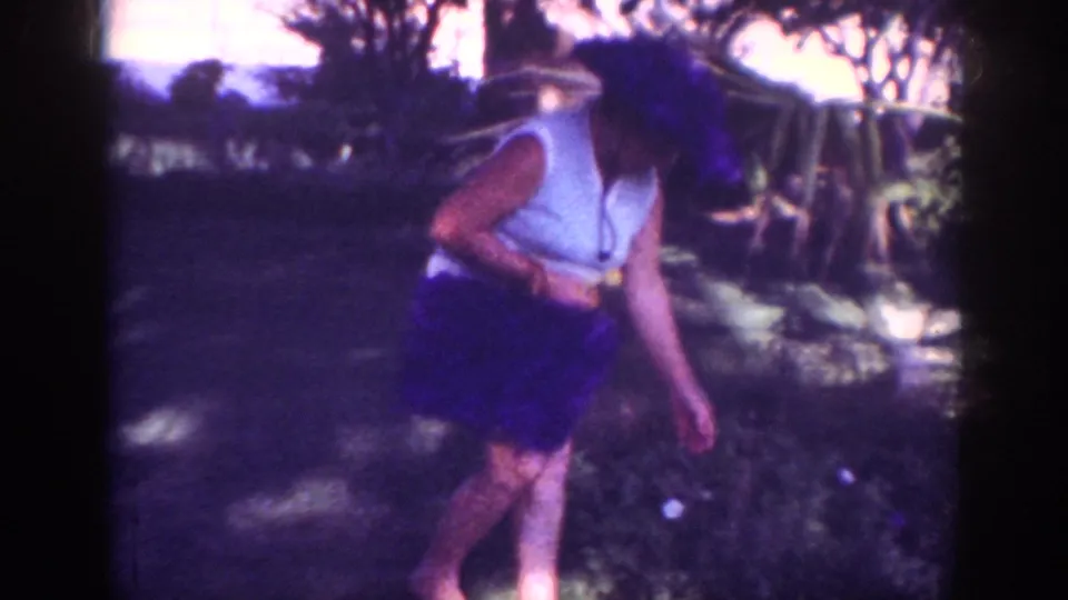 A 1962 Blainsport home-movie frame shows the woman leaning near garden shrubs with her handbag visible in the shade.