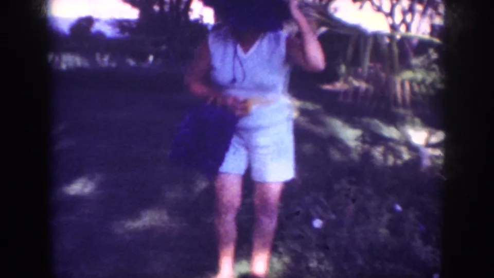 1962 Blainsport frame of a woman in a hat and pale outfit adjusting her headwear beside garden shrubs.