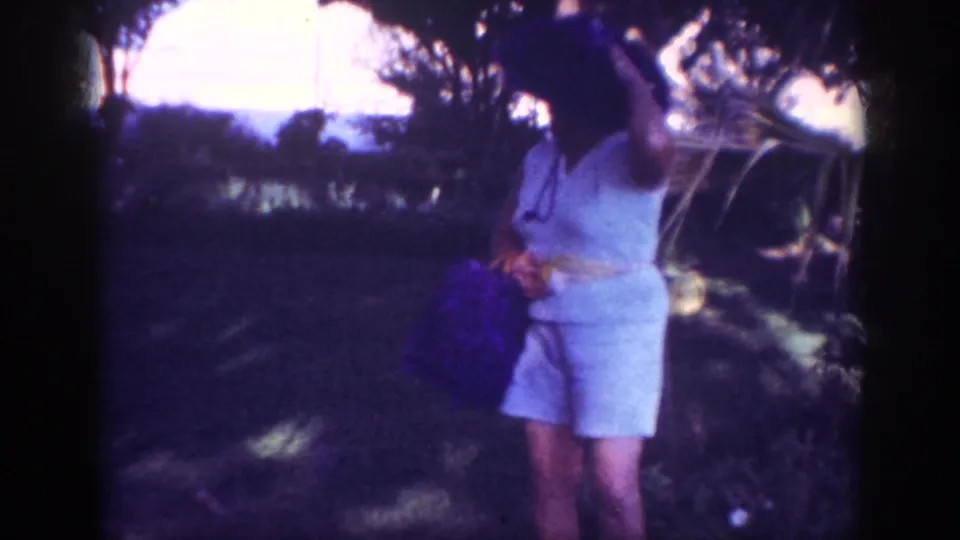 In Blainsport, Pennsylvania, 1962 home-movie footage, a woman in light clothing and a wide-brimmed hat stands in shaded grass near trees.