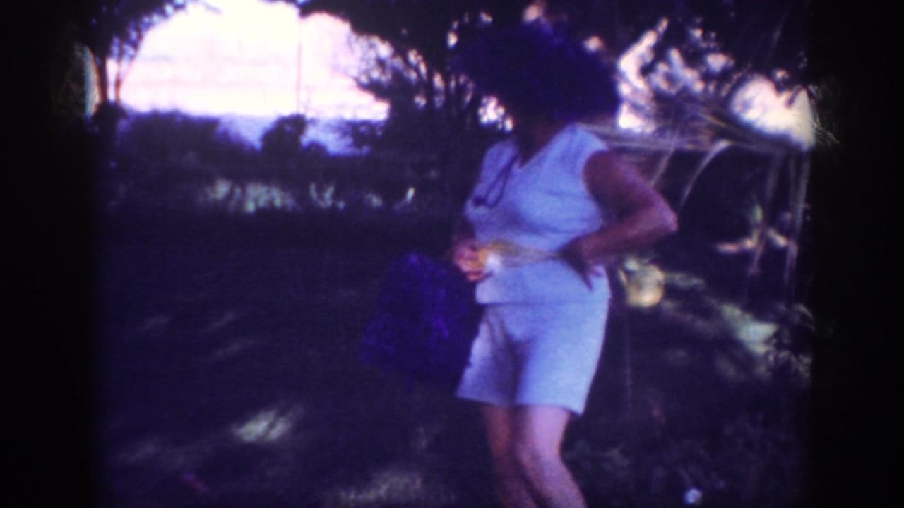 The woman stands under trees in the 1962 Blainsport picnic scene, holding a purple bag while shifting her pose for the camera.