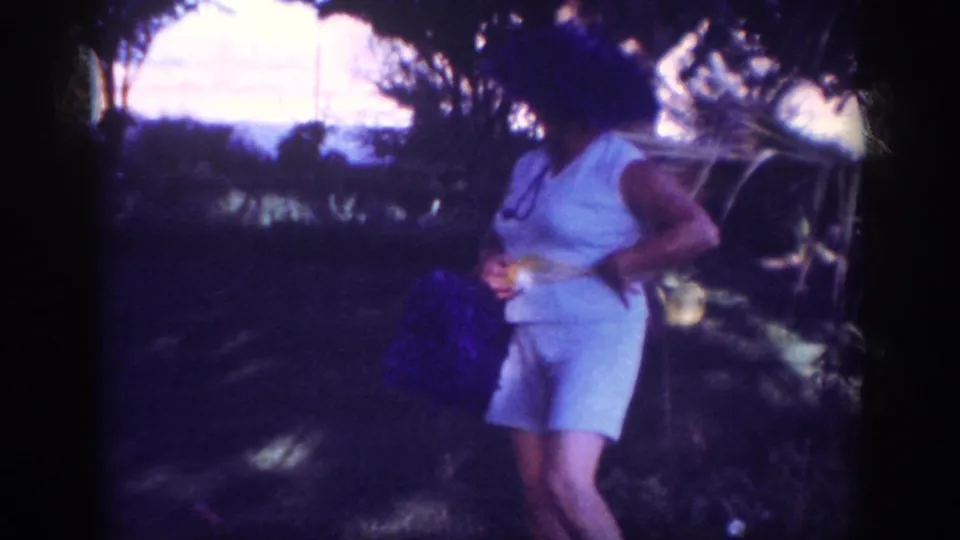 The woman stands under trees in the 1962 Blainsport picnic scene, holding a purple bag while shifting her pose for the camera.
