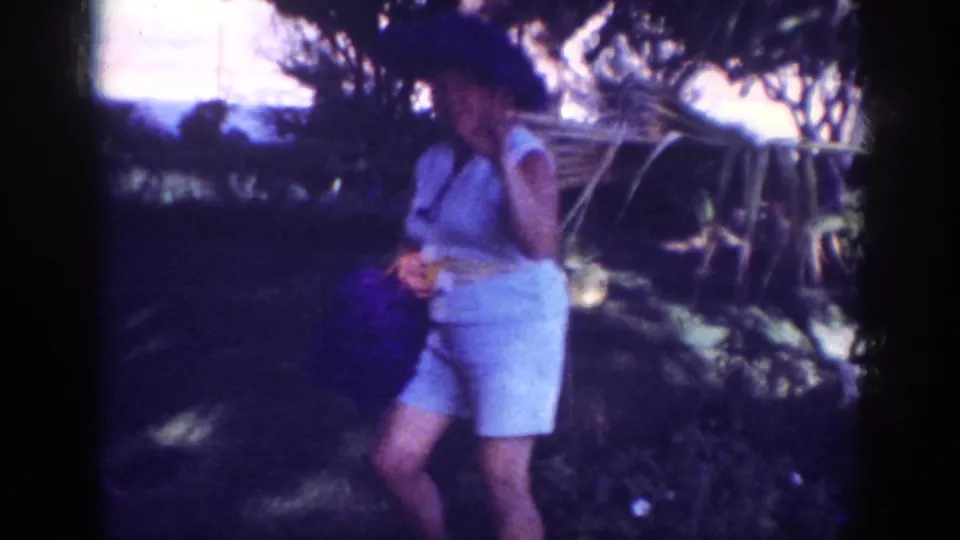 A woman in light clothing and hat moves through a shaded grassy area in the Blainsport 1962 home-movie picnic footage.