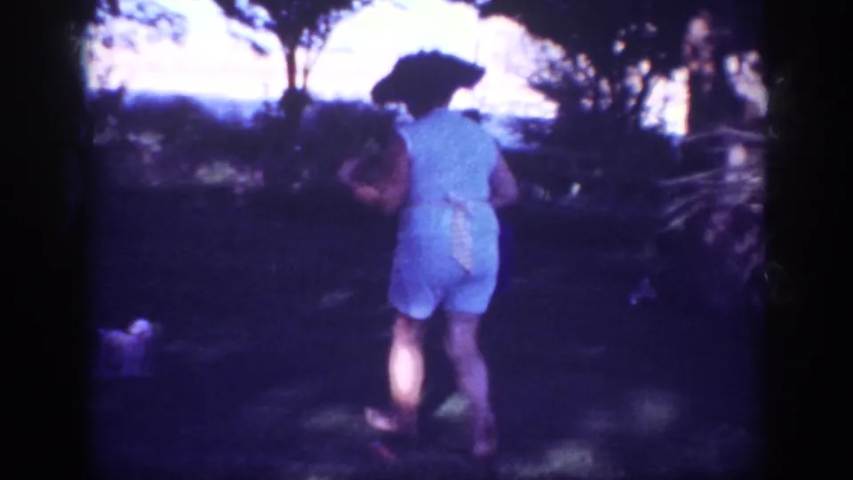 The woman walks away across shaded grass in the 1962 Blainsport footage, while a small dog appears near the lower left of the frame.