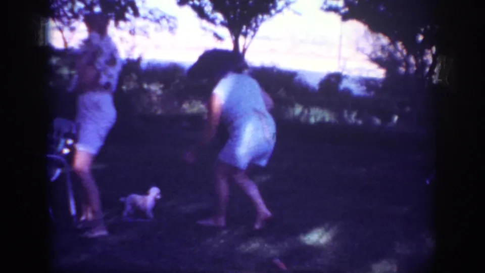 In Blainsport, Pennsylvania, 1962 home-movie footage, a woman in a light dress bends toward a small puppy in a shaded grassy yard.