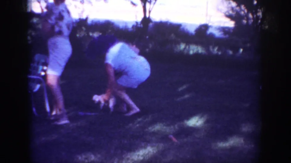 The woman bends low to catch or pick up the small puppy in a tree-lined yard, with another adult partly visible nearby.