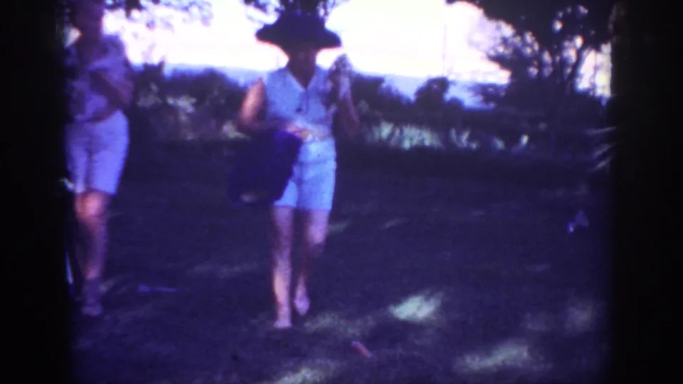 A woman wearing a wide-brimmed hat and light dress moves through the grassy yard in the 1962 Blainsport home-movie scene.