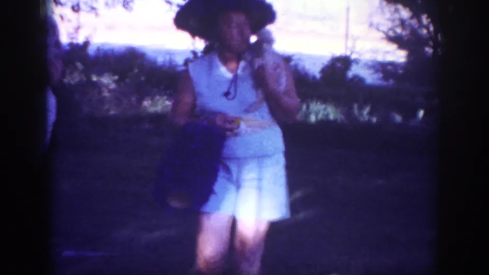 The woman stands in the yard holding the small puppy close, framed by trees and soft 8mm color from the Blainsport footage.