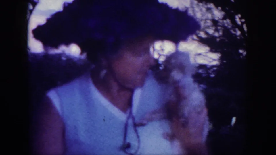 The woman, filmed in soft 8mm color, holds the small white dog near her face while standing among trees or shrubs.