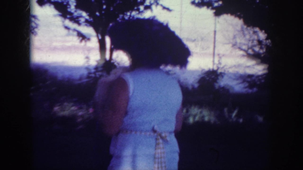 Woman in a pale dress and hat moving through the yard near trees and bright sky.