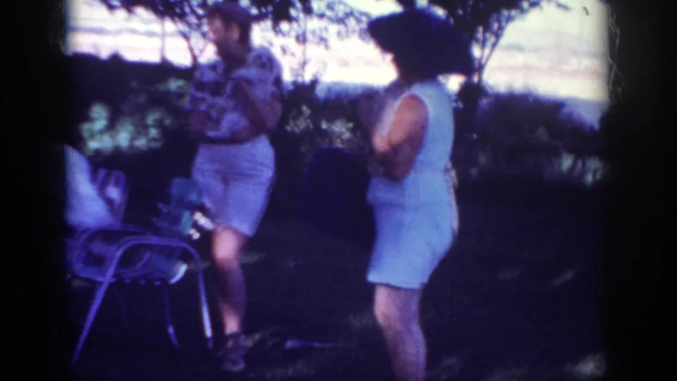 1962 Blainsport frame showing a woman in a pale dress near a man in patterned clothing, with metal patio chairs in the shaded backyard.