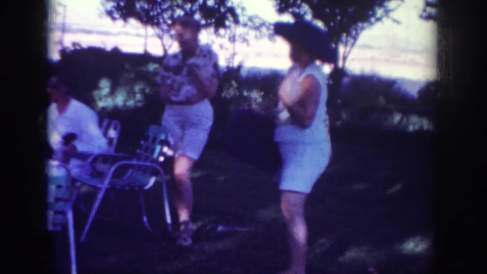 1962 Blainsport home-movie frame showing the woman beside a standing man, with patio chairs and a partly visible seated person nearby.