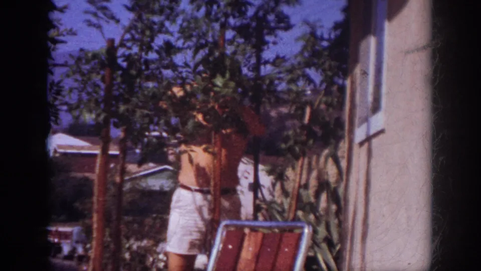 A shirtless man continues trimming a leafy tree near the house wall, with a patio chair and dense yard plantings visible.