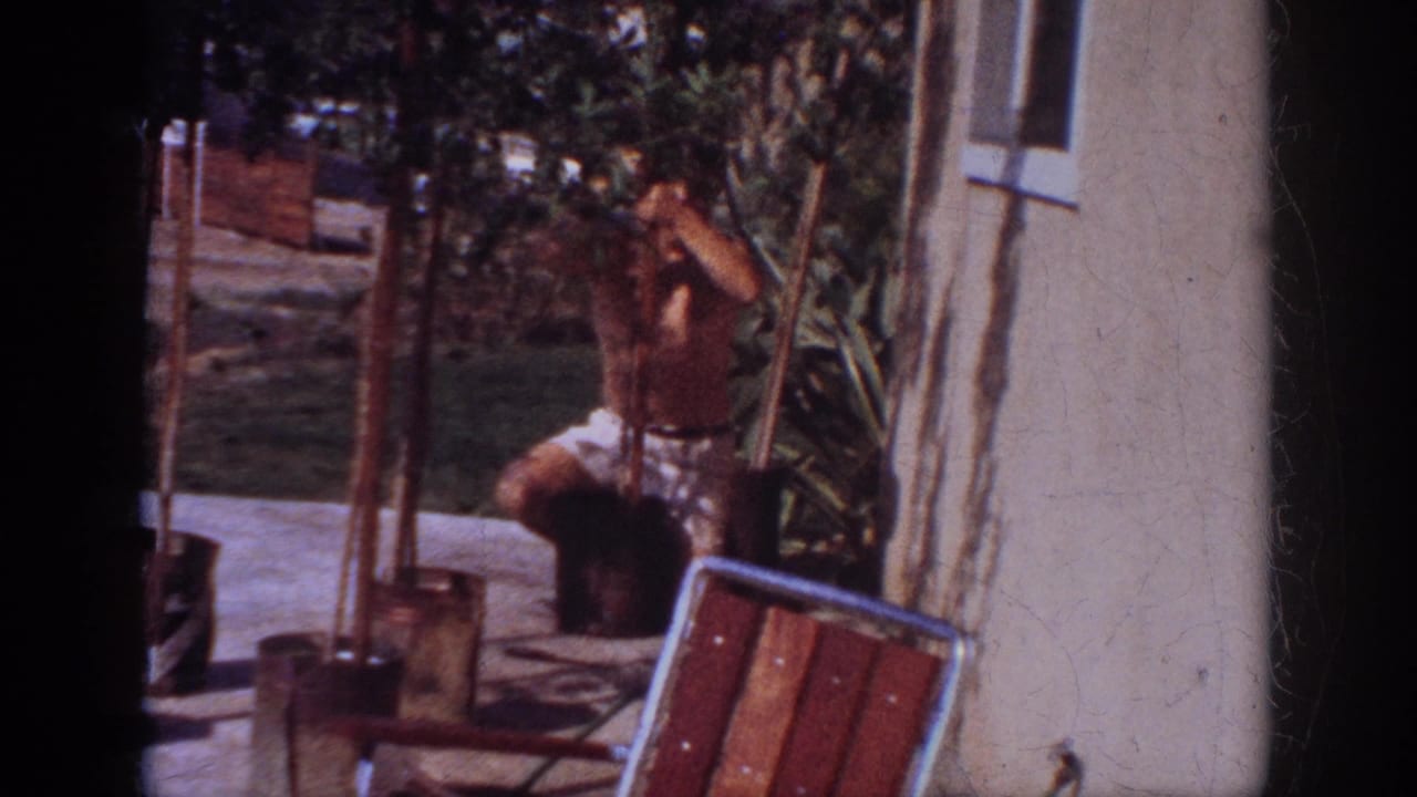 Man reaches toward a potted young tree near a chair and house wall.