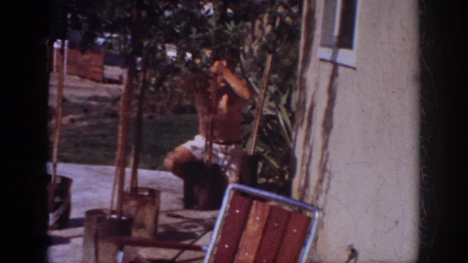 A shirtless man reaches into a young potted tree beside a patio chair and light house wall in the Blainsport backyard scene.