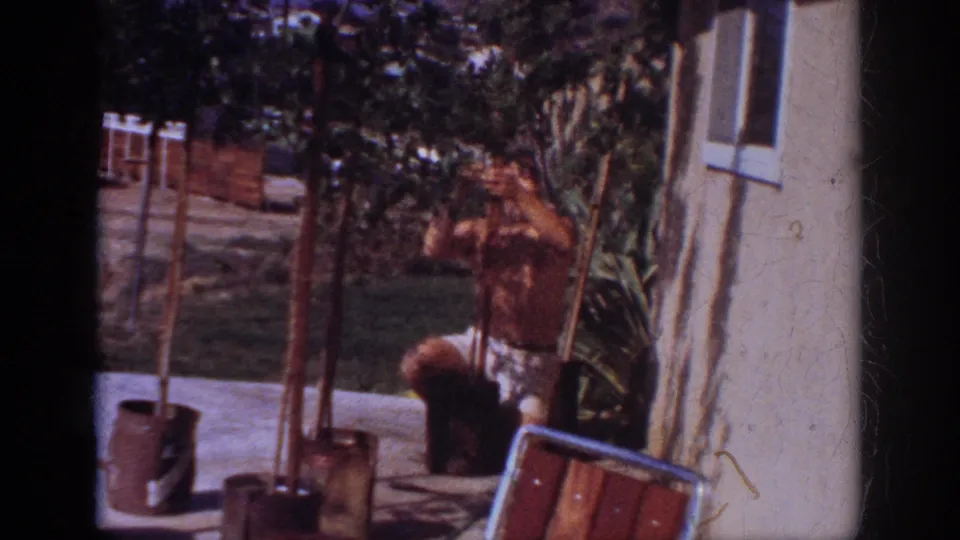 The man continues tending potted saplings beside the house, with a chair, fence, and sunlit yard visible around him.
