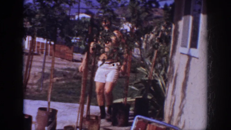 A wider frame shows the man among young trees and garden plants beside the house, with fence and yard details behind him.