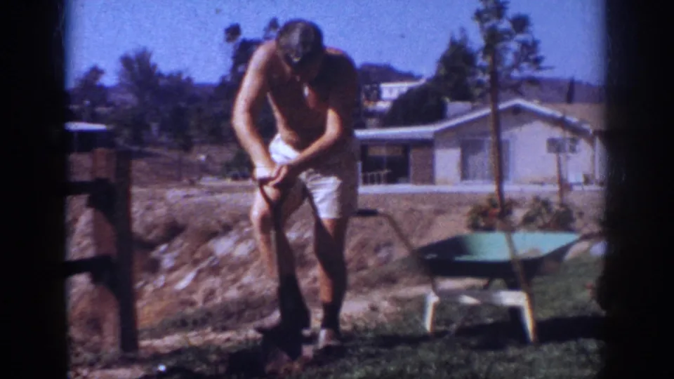 In a 1962 Pennsylvania backyard, the man bends over a shovel near bare soil, lawn, small trees, and a wheelbarrow.