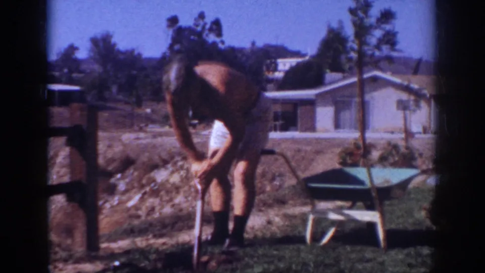 The man continues digging or planting near a wheelbarrow, with a single-story house and young trees behind him.