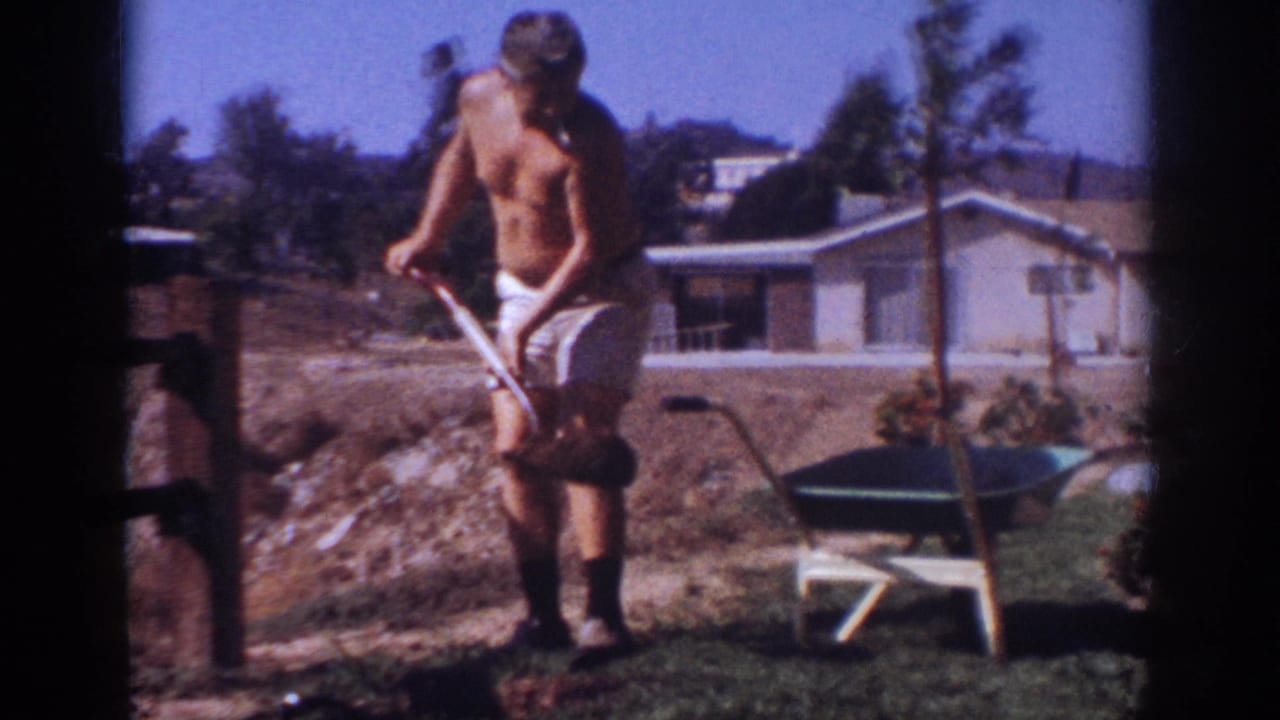 Man stands with a shovel near a young tree and wheelbarrow, with a house behind him.