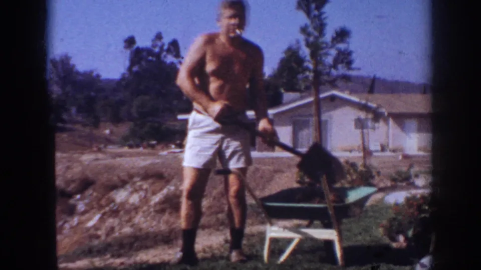 The man shovels soil near a green wheelbarrow, with a lawn, planted area, and single-story house visible in the background.