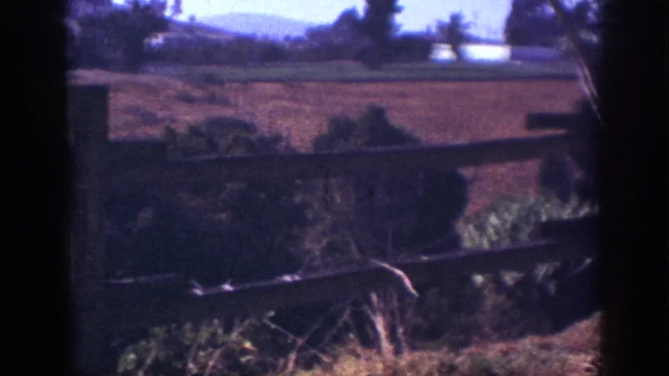 1962 Blainsport countryside frame showing fence rails, scrubby vegetation, open fields, and distant wooded hills.