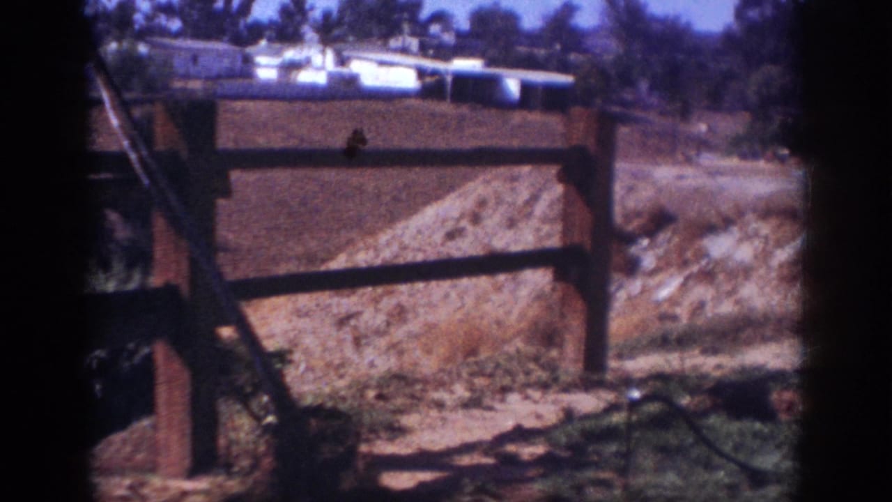 1962 Blainsport home-movie frame showing a wooden fence, dirt mound, sparse vegetation, and distant low buildings.