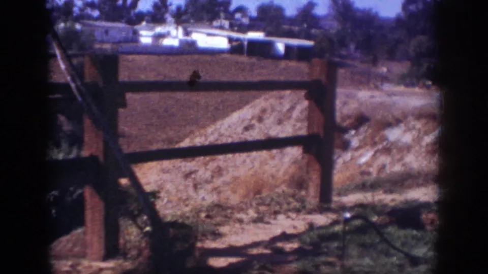 1962 Blainsport home-movie frame showing a wooden fence, dirt mound, sparse vegetation, and distant low buildings.