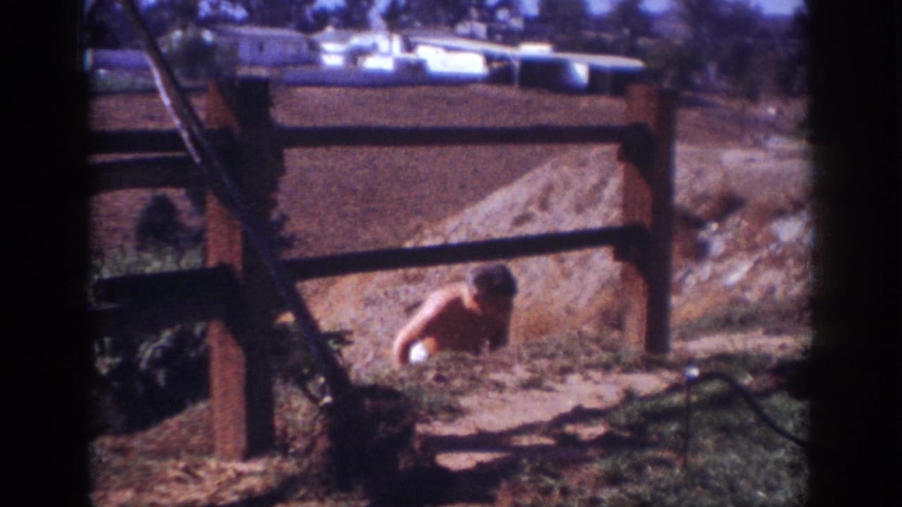 1962 Blainsport home-movie frame of a shirtless man crouching beside a dirt work area and wooden fence, with houses and trees beyond.