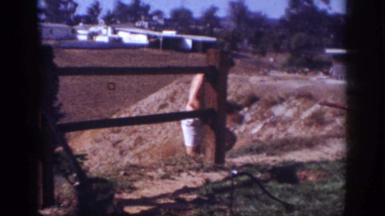1962 Blainsport frame showing a shirtless man in white shorts standing near a wooden fence and dirt mound in a residential yard.