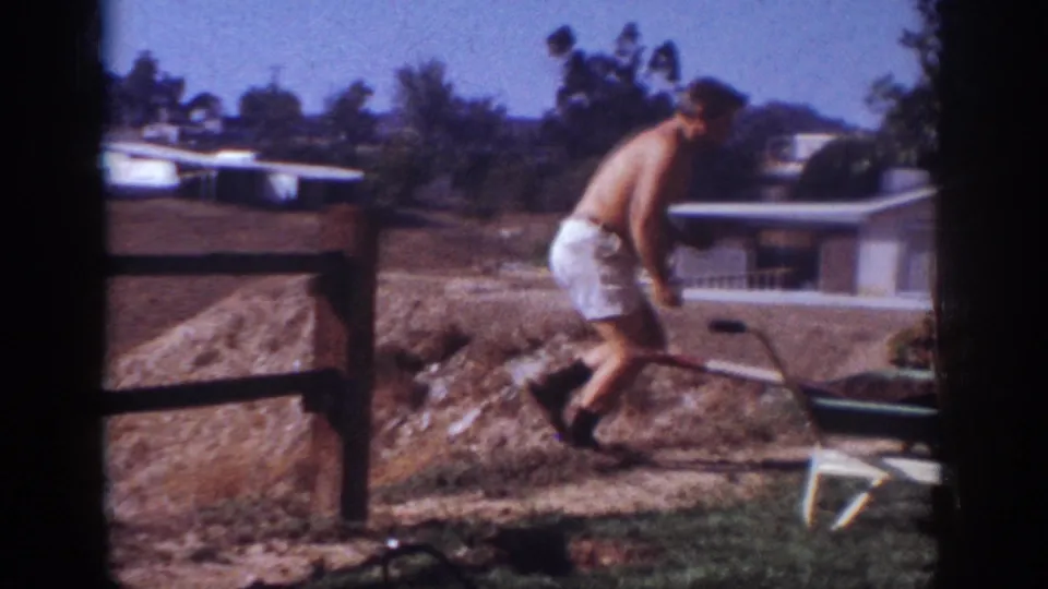 1962 Blainsport home-movie frame of the man moving near a wheelbarrow, with dirt piles, fence posts, house, and trees in the background.