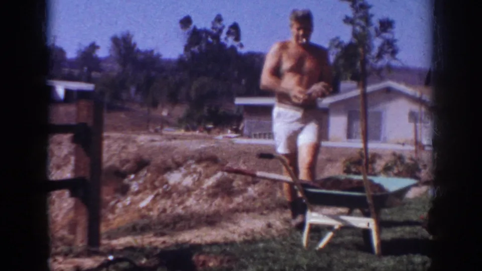 1962 Blainsport frame showing the man near a wheelbarrow in a residential yard, with a low house, trees, dirt, and grass visible.