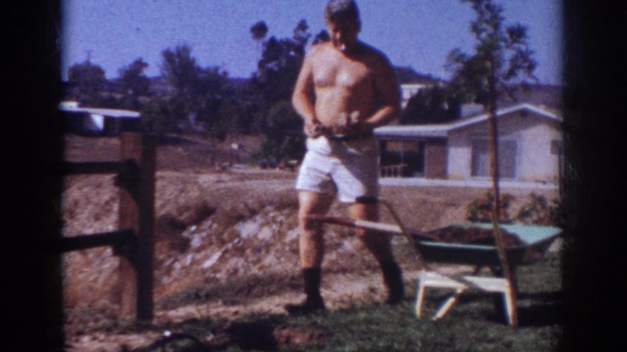 In 1962 Blainsport, Pennsylvania footage, a shirtless adult man stands in a suburban yard beside a wheelbarrow and fence.