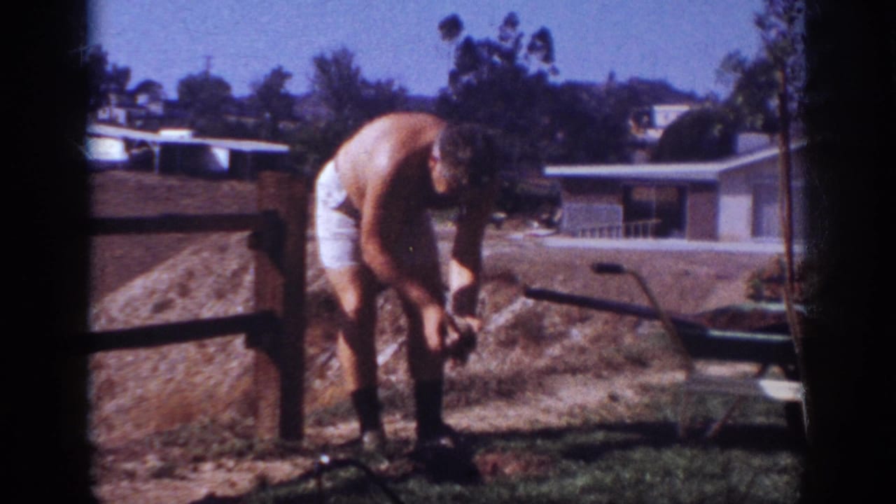 Shirtless man bends over a patch of soil with a wheelbarrow nearby.